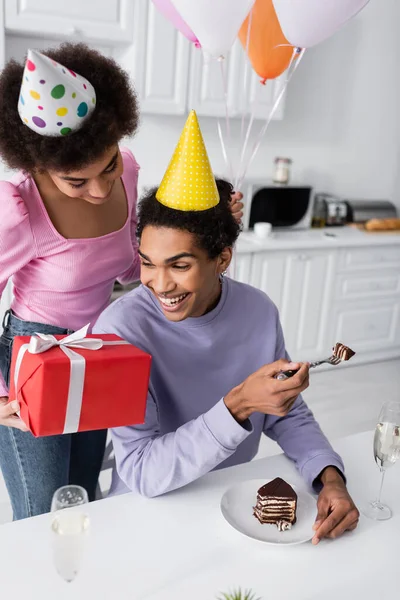 Homem americano africano positivo segurando bolo de aniversário perto da namorada com presente e balões na cozinha — Fotografia de Stock