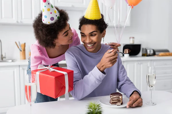 African american woman in party cap holding balloons and present near boyfriend with birthday cake and champagne in kitchen — Stockfoto