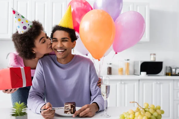 Африканская американка с воздушными шарами и подарками целует улыбающегося бойфренда с тортом на день рождения дома — стоковое фото