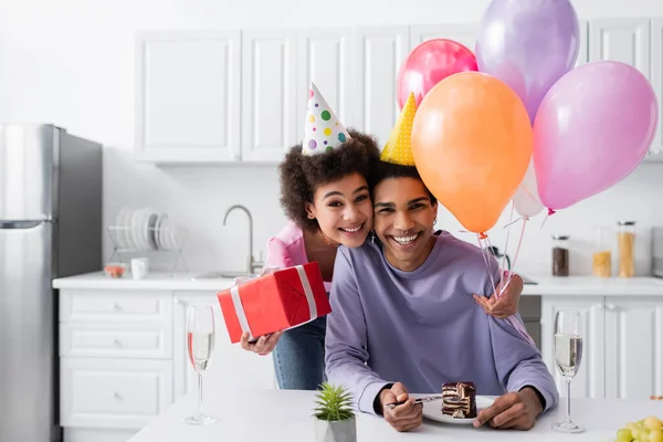 Heureuse femme afro-américaine en bonnet de fête tenant cadeau et ballons près du petit ami, gâteau d'anniversaire et champagne à la maison — Photo de stock