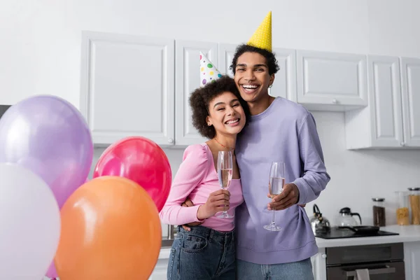 Heureux homme afro-américain en bonnet de fête tenant champagne et étreignant petite amie près de ballons dans la cuisine — Photo de stock