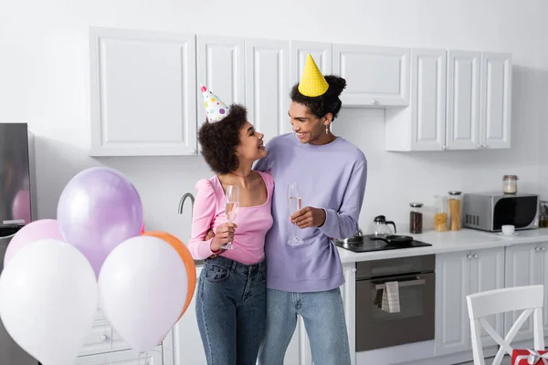 Positif homme afro-américain tenant un verre de champagne et étreignant petite amie dans le chapeau de fête près de ballons à la maison — Photo de stock