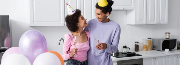 Cheerful african american man in party cap holding champagne and hugging girlfriend near balloons at home, banner — Stock Photo