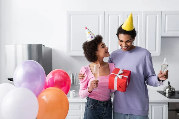 Smiling african american woman in party cap holding gift and champagne near boyfriend and balloons in kitchen — Stock Photo