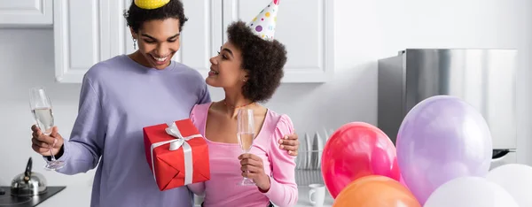 Joven mujer afroamericana en gorra de fiesta sosteniendo champán y caja de regalo cerca de novio y globos en casa, pancarta - foto de stock