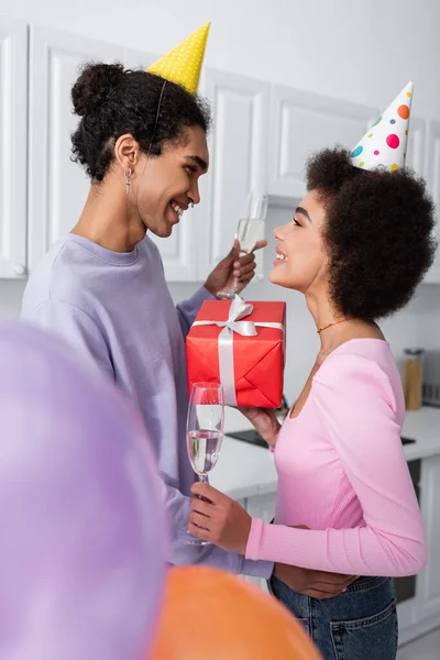 Vue latérale de souriant homme afro-américain en chapeau de fête tenant champagne et étreignant petite amie avec présent près de ballons à la maison — Photo de stock