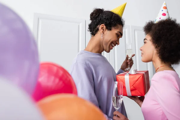 Vista laterale della donna afro-americana con champagne e regalo vicino al fidanzato in berretto da festa e palloncini sfocati a casa — Foto stock
