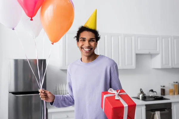 Happy african american man in party cap holding gift and balloons while looking at camera at home — Stock Photo