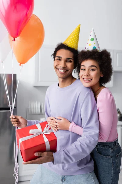 Mulher americana africana alegre em boné de festa abraçando namorado com balões e presente em casa — Fotografia de Stock