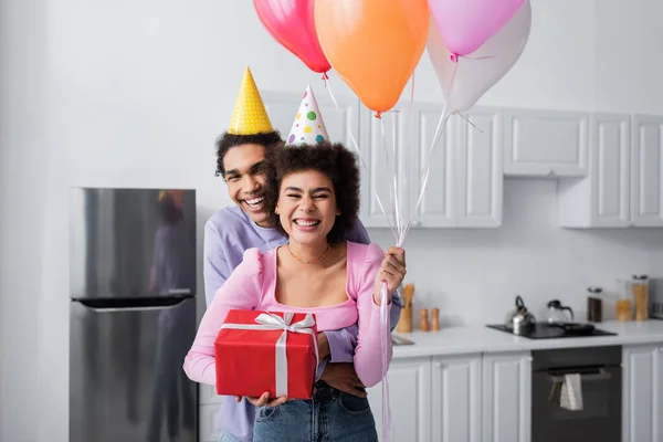 Joven afroamericano hombre en partido gorra abrazando novia con presente y globos en casa - foto de stock