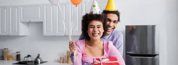 Позитивний афроамериканець на вечірці обнімає дівчину з сувенірною коробкою і кульками вдома, банер. — стокове фото