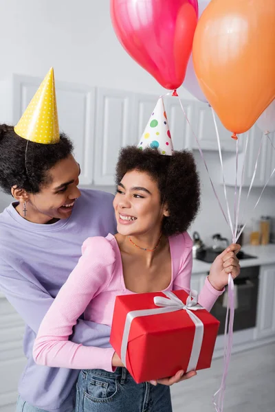 Sonriente hombre afroamericano en gorra de fiesta abrazando novia con globos festivos y regalo en casa - foto de stock