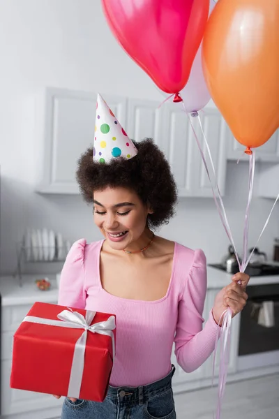 Joven afroamericana mujer en partido gorra celebración regalo y globos en casa - foto de stock