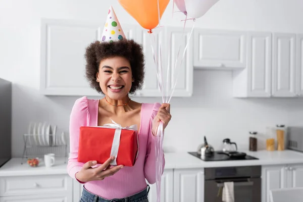 Cheerful african american woman in party cap holding gift box and balloons at home — Fotografia de Stock