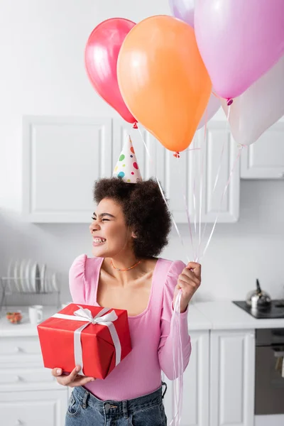 Happy african american woman in party cap holding gift and balloons in kitchen at home — Fotografia de Stock