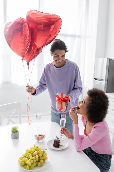 Smiling african american man holding heart-shaped balloons and gift near girlfriend with strawberry and champagne at home — Foto stock
