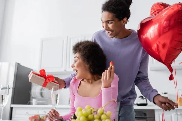 Happy african american man holding heart-shaped balloons and gift near girlfriend with present and strawberry at home — Foto stock