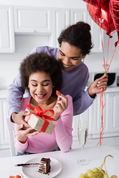 Smiling african american man holding balloons and gift near girlfriend with strawberry and champagne at home — Foto stock