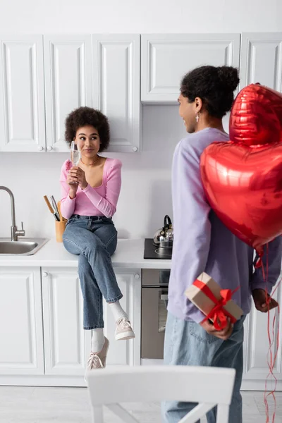 Hombre afroamericano escondiendo globos en forma de corazón y presente cerca de novia con champán en casa - foto de stock