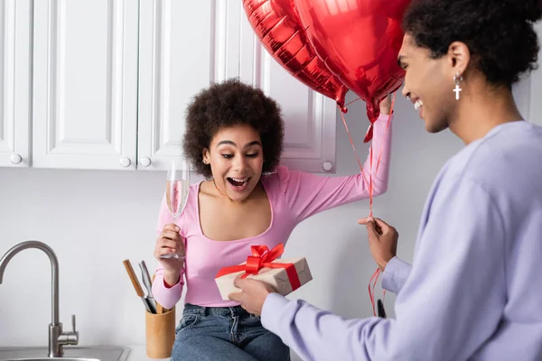 Smiling african american man holding balloons and gift near excited girlfriend with champagne in kitchen — Foto stock