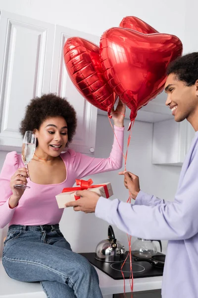 Sonriente mujer afroamericana sosteniendo champán cerca de novio con globos en forma de corazón y caja de regalo en la cocina - foto de stock