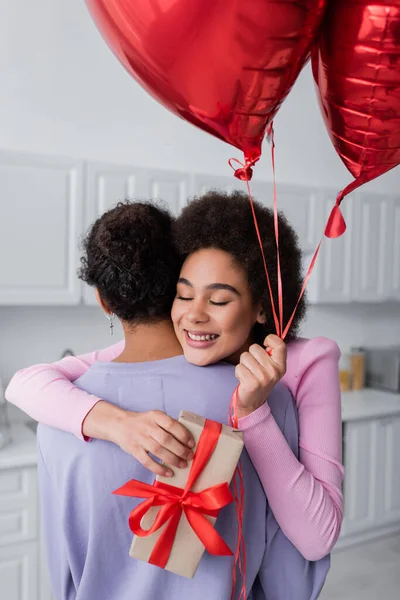 Молодая африканская американка держит воздушные шары в форме сердца и подарок, обнимая своего парня дома — стоковое фото