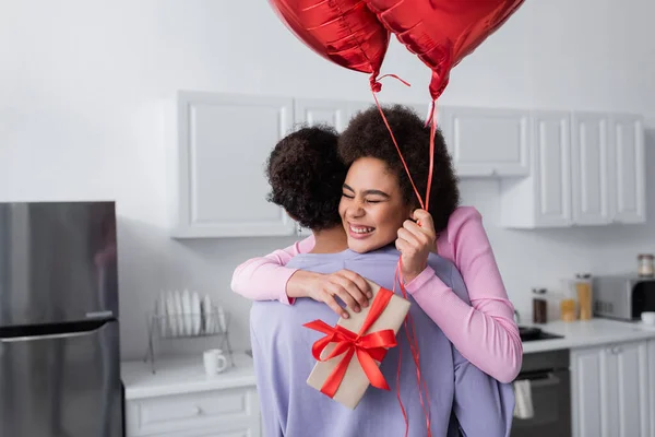 Веселая африканская американка держит воздушные шары и подарок, обнимая своего парня дома. — стоковое фото