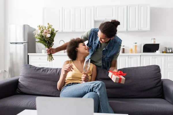 Afroamerikaner hält Blumenstrauß und Geschenk in der Nähe von Freundin mit Champagner zu Hause — Stockfoto