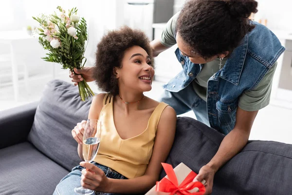 Uomo afroamericano in possesso di bouquet e presente vicino sorridente fidanzata con champagne a casa — Foto stock