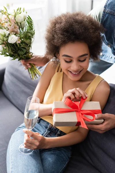 Smiling african american woman holding champagne near boyfriend with gift and flowers at home — Stock Photo