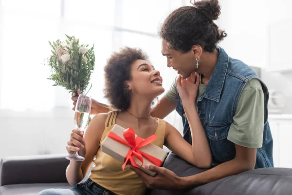 Femme positive tenant champagne près copain afro-américain avec cadeau et bouquet à la maison — Photo de stock