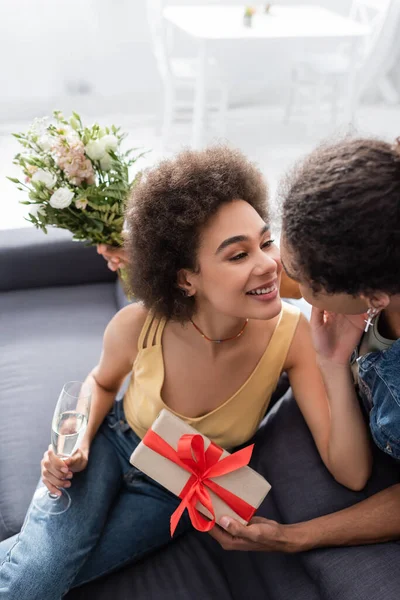 Vista ad alto angolo della donna afro-americana sorridente che tiene champagne vicino al fidanzato con fiori e scatola regalo a casa — Foto stock