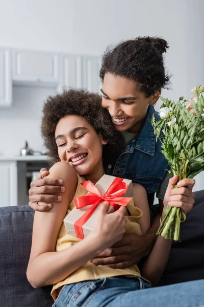 Jovem afro-americano homem abraçando namorada com flores e presente em casa — Fotografia de Stock