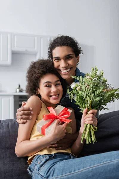 Smiling african american couple with bouquet and present looking at camera on couch at home — Foto stock