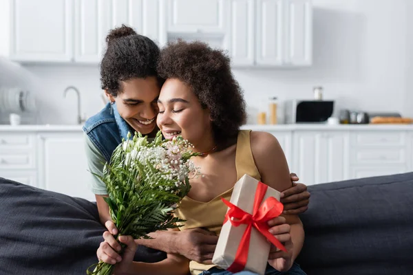 Jovem afro-americano homem abraçando sorridente namorada com buquê e presente em casa — Fotografia de Stock