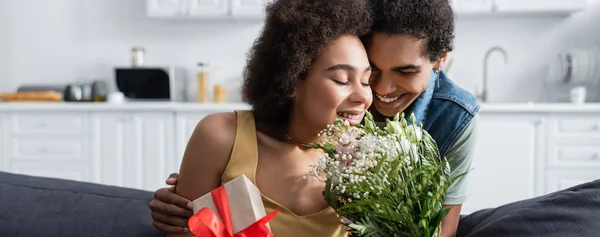 Glücklicher afrikanisch-amerikanischer Mann umarmt Freundin mit Geschenk und Blumen zu Hause, Banner — Stockfoto