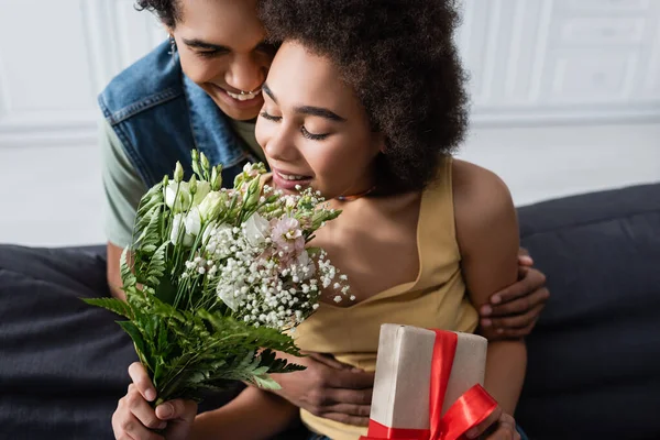 Sorrindo afro-americano homem abraçando namorada com buquê e presente em casa — Fotografia de Stock
