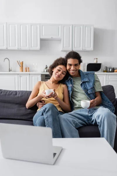 Souriant homme afro-américain tenant tasse et étreignant petite amie près d'un ordinateur portable flou à la maison — Photo de stock
