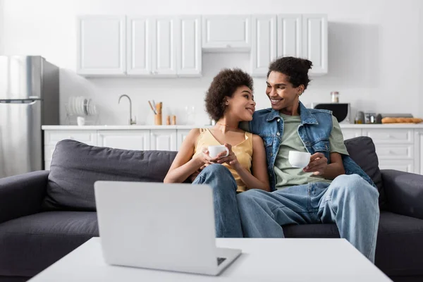 Joven pareja afroamericana sosteniendo tazas cerca de la computadora portátil en casa - foto de stock