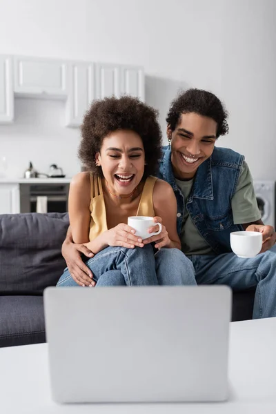 Alegre pareja afroamericana con tazas mirando borrosa portátil en casa - foto de stock
