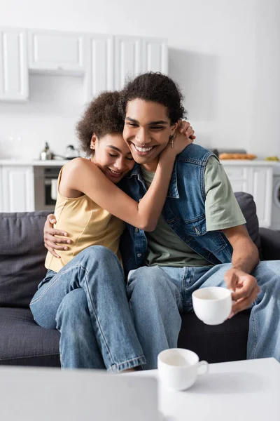 Cheerful african american woman hugging boyfriend with cup near laptop at home — Foto stock