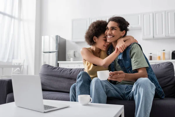 Young african american woman with closed eyes hugging smiling boyfriend with cup near laptop at home — Fotografia de Stock