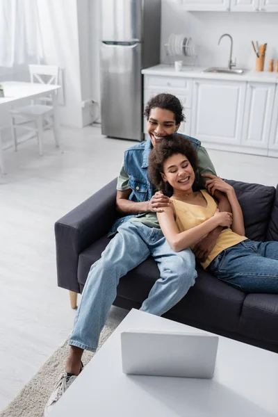 Africano americano hombre abrazando sonriente novia y mirando portátil en casa - foto de stock