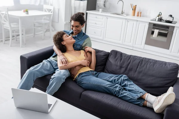 Jovem casal afro-americano abraçando no sofá perto de laptop em casa — Fotografia de Stock