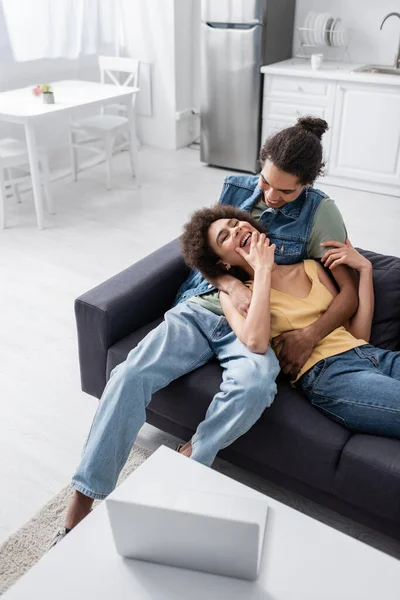 Позитивний афроамериканець, який обіймає веселу дівчину на дивані біля ноутбука вдома. — стокове фото