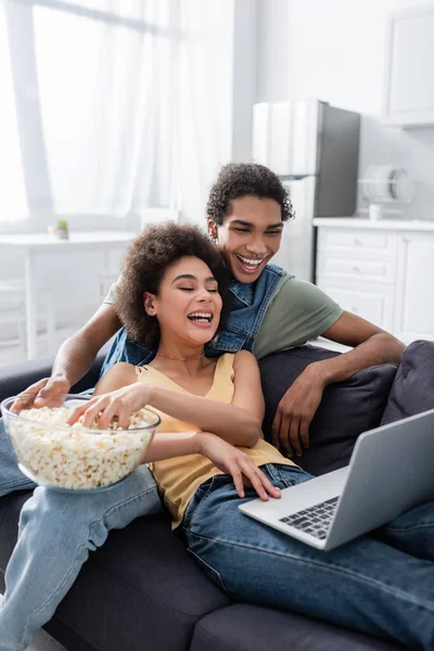 Улыбающаяся африканская американская пара держит попкорн, глядя на ноутбук дома — стоковое фото