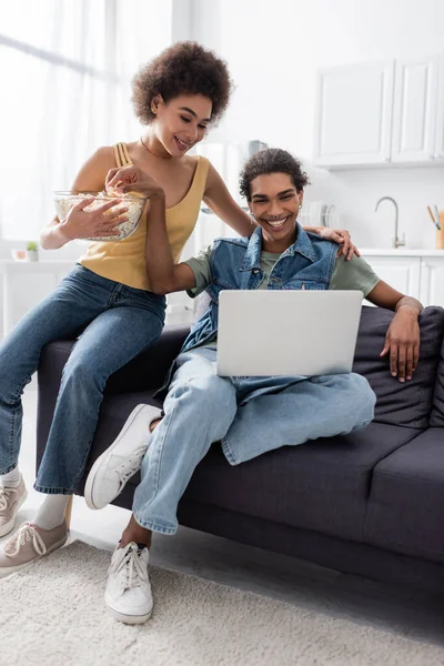 Femme afro-américaine tenant pop-corn près de petit ami avec ordinateur portable sur le canapé à la maison — Photo de stock