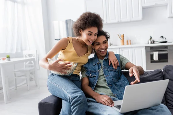 Lächelnde Afroamerikanerin hält Popcorn in der Hand und umarmt Freund zu Hause mit Laptop — Stockfoto