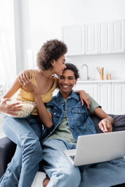 Joven mujer afroamericana abrazando novio y sosteniendo palomitas de maíz cerca del ordenador portátil en el sofá - foto de stock