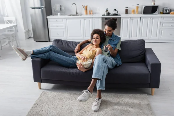 Souriant homme afro-américain tenant télécommande près de petite amie avec pop-corn à la maison — Photo de stock